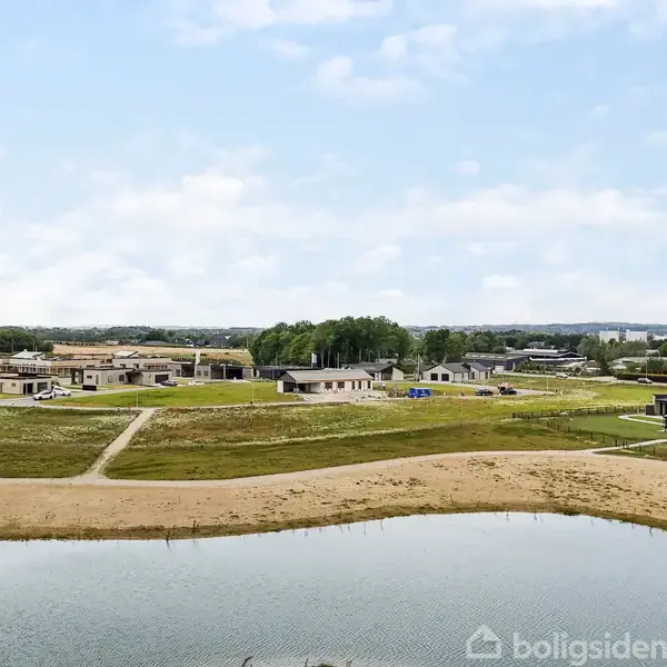 Flere moderne enfamiliehuse ligger spredt langs velordnede grusstier i et åbent, nybygget boligområde med en sø i forgrunden og skov i baggrunden under en klar blå himmel.