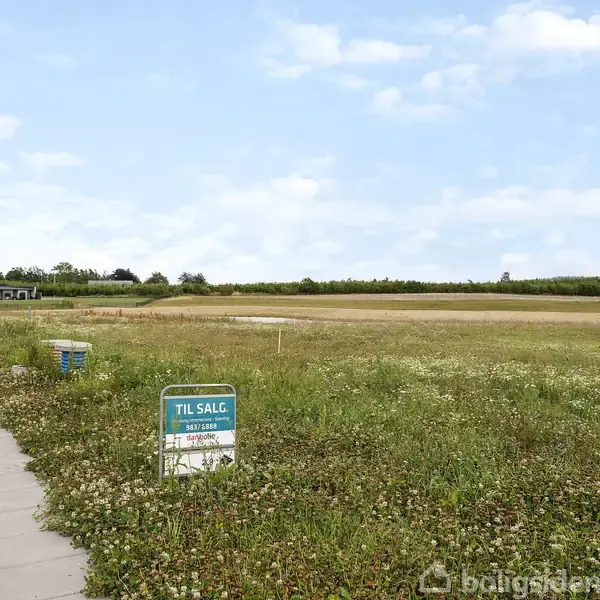 Græsmark med skilt "TIL SALG", kontaktoplysninger for mægler "Bo Nygaard Larsen" og "Danbolig", ved siden af en flisebelagt sti og omgivet af marker.