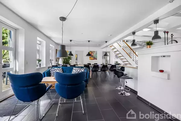 Blå lænestole står omkring et træbord i en moderne, lys stue med sort klinkegulv. I baggrunden ses en spiseplads med sorte stole og et åbent køkkenområde under en trappe.