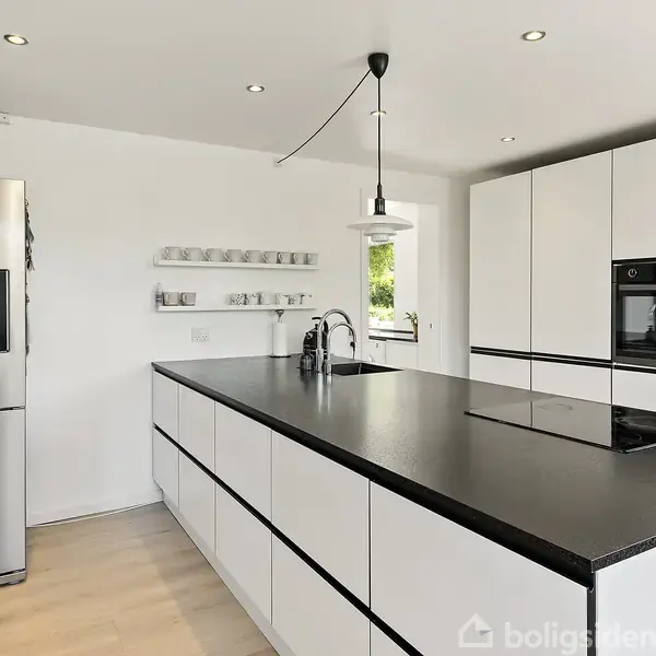 Et moderne køkken med en stor, sort køkkenø i midten, indbyggede ovne i skabsvæggen og et køleskab til venstre. Hvide vægge og trægulv skaber en lys atmosfære.