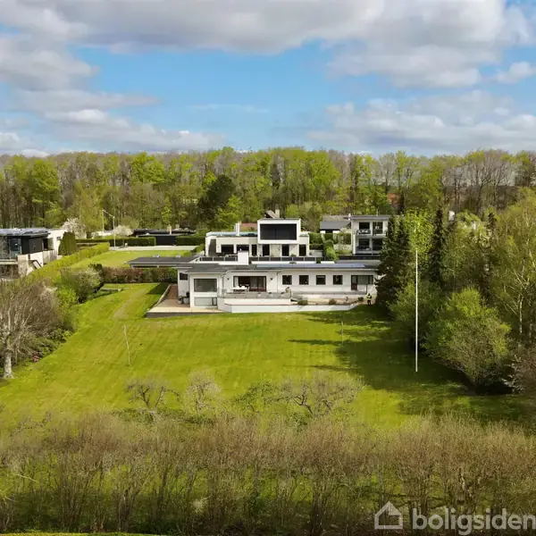 En moderne, hvid villa står omgivet af en stor, velplejet græsplæne med træer; placeret i en rolig, skovklædt forstadsmiljø under en blå, let skyet himmel.