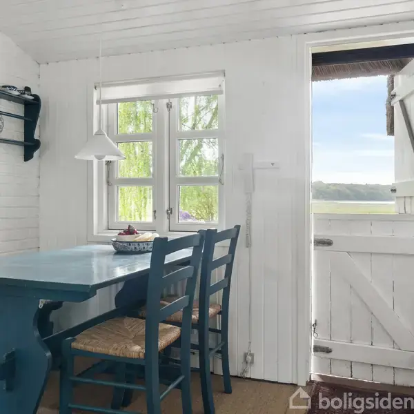 Blåt spisebord med tre stole står i en lille, hvidmalet spisestue. En åben stalddør fører til en udsigt over marklandskab udenfor. En lampe hænger over bordet.