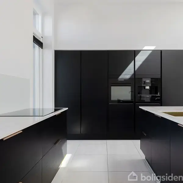 Moderne køkken med sort skabsfront, indbygget ovn og kogeplade. En hvid bordplade og messinghane står overfor store sorte skabe. Lyse vægge og gulvfliser skaber en minimalistisk atmosfære.