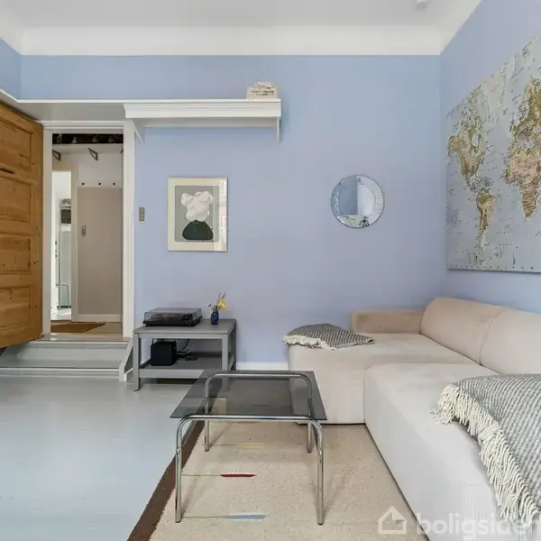 En lys stue med en stor, beige hjørnesofa og et glasbord i midten. En verdenskortplakat hænger på en blå væg. Et åbent døråbning fører til en gang. Ingen tekst fundet.