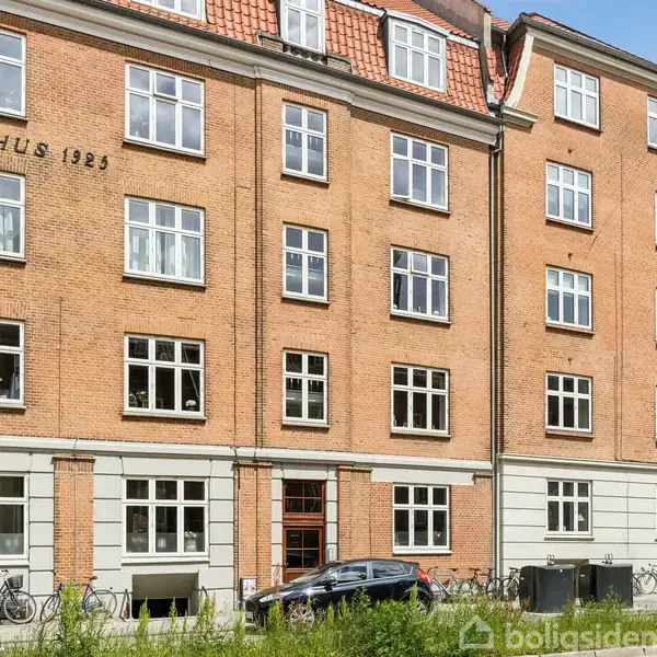Bygning med navnet "HAMMELHUS 1923" i røde mursten med hvidrammede vinduer. Foran bygningen holder biler parkeret langs vejen, og der ses cykler på fortovet.