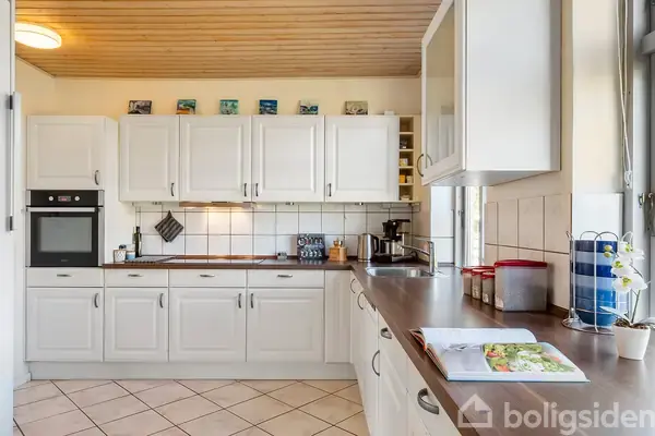 Et køkken med hvide skabe, en ovn og kogeplade i aktion. Konteksten er et lyst rum med fliser på væggen og bordet. En åben bog ligger på bordet.