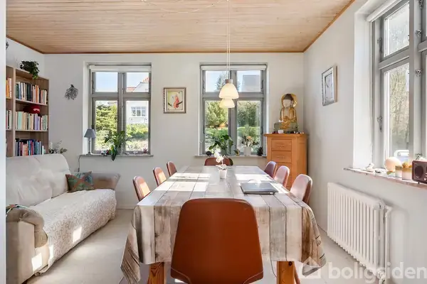 Spisebord med dug og seks stole står i en lys stue. Til venstre er en sofa og bogreol, mens der til højre er flere vinduer med udsigt til grønne omgivelser.