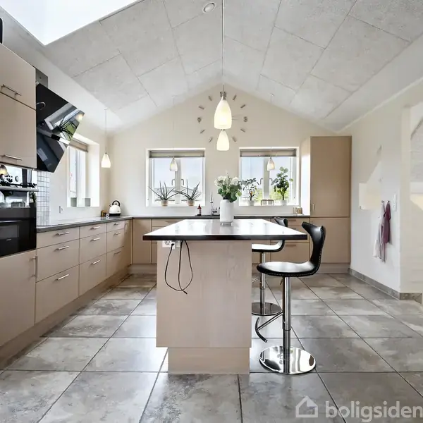 Et moderne køkken med lyse træskabe og en central ø med barstol. Hængende lamper lyser rummet, der har fliser på gulvet og store vinduer med planter.