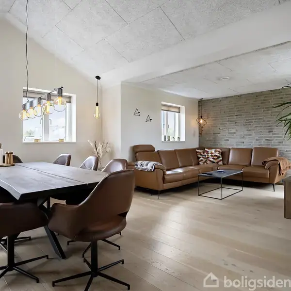 Spisebord med brune stole står i en moderne stue med en hjørnesofa og sofabord. Et væghængt TV og grønne planter tilføjer til indretningen. Loftslamper lyser rummet.