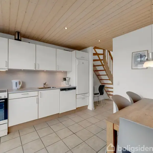 Køkken med hvide skabe og apparater inklusive komfur og elkedel. Træloft og flisegulv. Et spisebord med stole står nær en trappe, der fører til en anden etage.