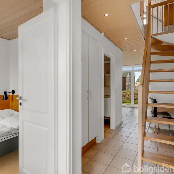 En trappe fører op til øverste etage i et hus med lyse vægge og lofter. Til venstre er en åben dør til et soveværelse med seng og lampe.