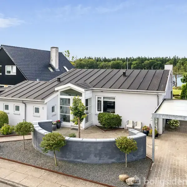 Et hvidt hus med mørkt tag står stille omgivet af en velplejet have. I baggrunden ses en sø og skov. Foran huset er en indkørsel med en carport.