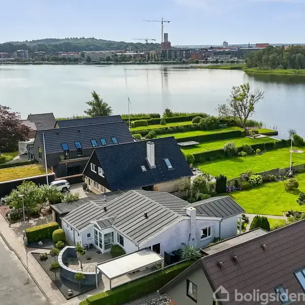 Et hvidt hus med sort tag står i en velplejet have. Baggrunden viser en sø og en by med bygninger og en kran i horisonten.