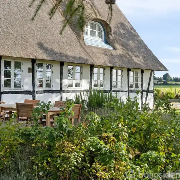 Et bindingsværkshus med stråtag står i en have med buske og træer. Udendørs træmøbler er placeret nær huset. Landskabet i baggrunden viser marker under en blå himmel.