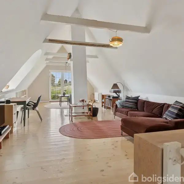 Stue med skrå vægge, hvor en brun sofa står på et trægulv. Et lille bord og stole er placeret ved en vinduesfront. Lyse træmøbler og rødligt tæppe.