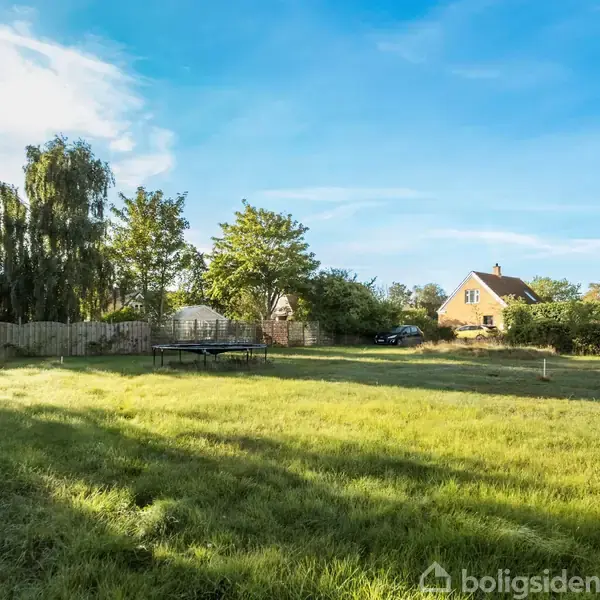 En græsplæne ligger stille i sollys, omgivet af et hegn og træer, med et hus i baggrunden under en klar blå himmel.