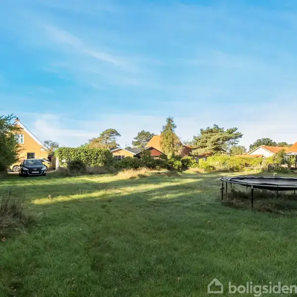 Trampolin står stille på en stor græsplæne i en åben have. I baggrunden ses huse, træer og en parkeret bil, alt under en blå himmel.