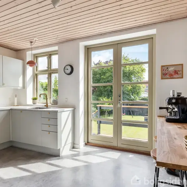 Et lyst køkken med hvide skabe og en kaffemaskine på en træbordplade. Store vinduer og glasdøre giver udsigt til en solrig have med frodige træer.