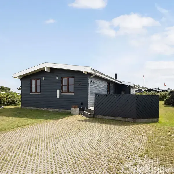 En étplans træhus med grå facade og hvidt tag ligger på en græsplæne omgivet af lave buske. Huset har vinduer med brune rammer, og himlen er delvist overskyet.