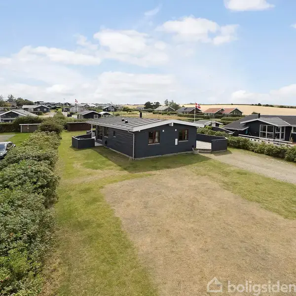 Et enkelt, mørkt sommerhus står på en stor, åben græsplæne omgivet af lave buske og andre huse i et villakvarter. Himlen er let skyet.