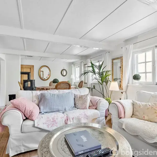 En lys stue med to hvide sofaer fyldt med farverige puder. En rund kaffebord med blade og bøger foran sofaerne. Vinduerne lader naturligt lys ind, omgivet af planter og vægkunst.