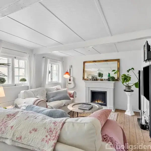 Stue med hvide vægge, loftsbjælker og gulve i træ. Sofaer arrangeret omkring et rundt sofabord. Kamin med spejl og planter. Vinduer med gardiner slipper lys ind.