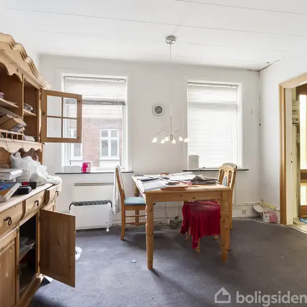 Reol med åbne skabe står ved væg. Bord og stole er dækket af papirer og stof. To vinduer slipper lys ind. Trægulv og dør i baggrunden.