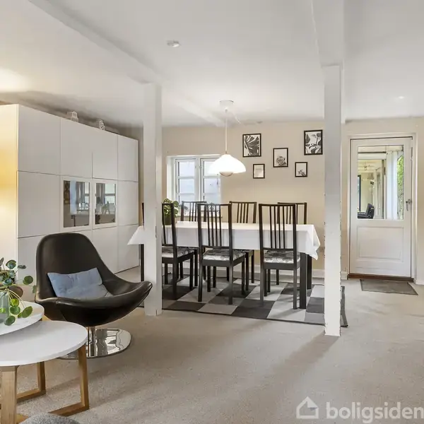 En stue med moderne indretning, hvor en sort lænestol og runde borde står ved siden af en sofa. Et spisebord med stole ses i baggrunden under en lampe.