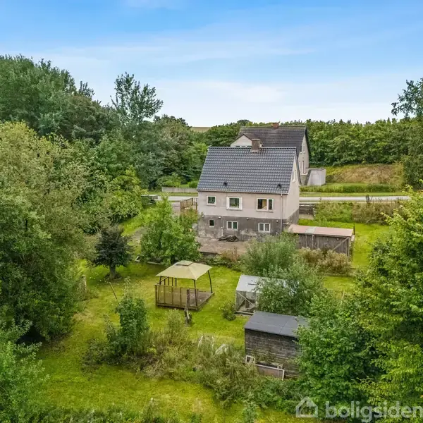 Hus står stille omgivet af frodige træer og buske i en grøn have. Flere små skure og legeplads er synlige i forgrunden, med vej og skov i baggrunden.