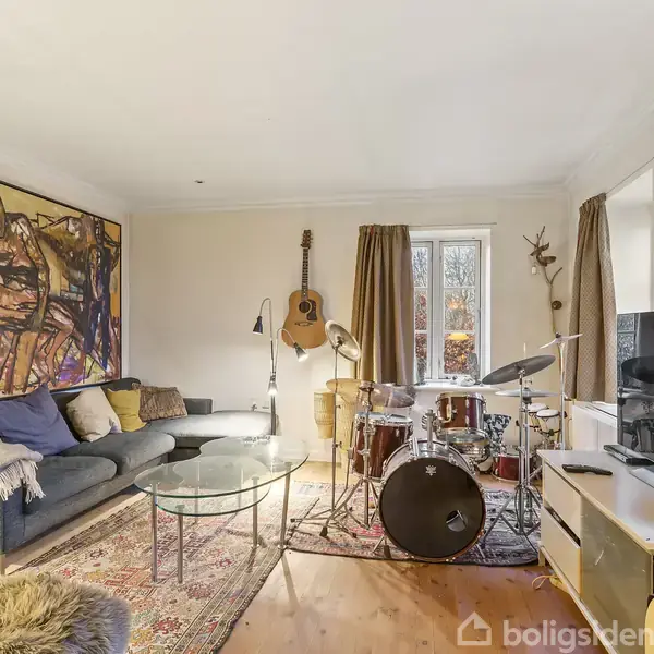 Et moderne stueinteriør med et trommesæt placeret nær et vindue. Omkring står en sofa, guitarer, et stort farverigt maleri og et fjernsyn. Tæpper dækker det trægulv.