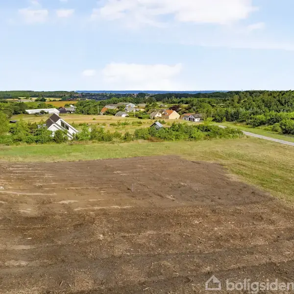 Tomt jordstykke ligger forrest, omgivet af en grusvej til højre og spredte huse i baggrunden. Landskabet er frodigt med grønne skove og åbne marker under en blå himmel.