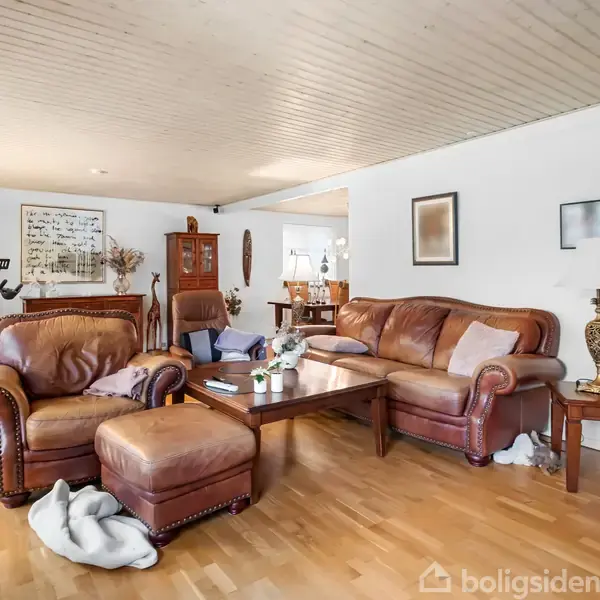Sofagruppe i brunt læder står i en stue med trægulve, lysloft og hvide vægge. Rummet indeholder lamper, et sofabord og pyntegenstande. Et træskab og motionscykel ses i baggrunden.