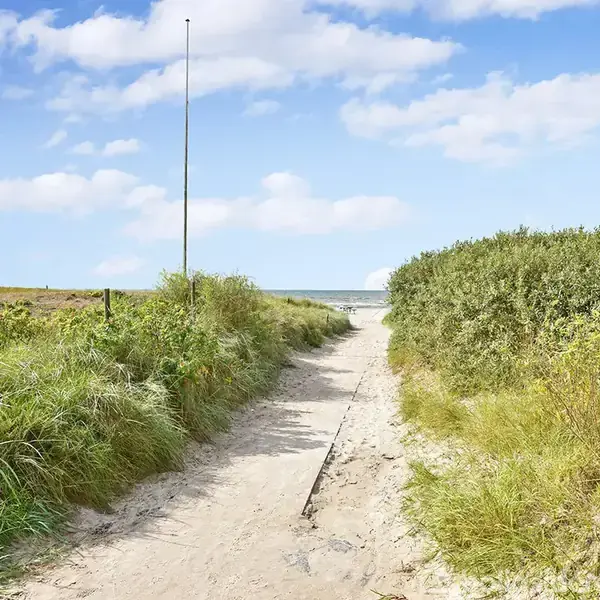 Sti strækker sig gennem sand og græs, fører mod stranden. Informationsskilt står til højre. Blå himmel med spredte skyer ses i baggrunden.