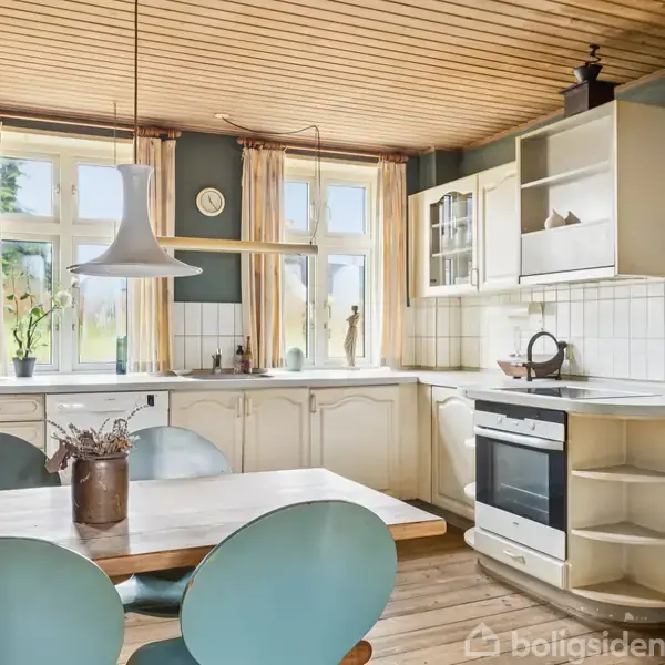 Et køkkenbord med blå stole står foran en L-formet køkkenbordplade med integreret ovn og skabsløsninger. En lampe hænger over bordet. Store vinduer giver naturligt lys.