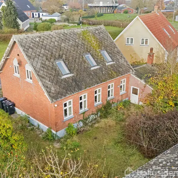 Rødt murstenshus med skråt tag og ovenlysvinduer, placeret i en have med buske. Naboer og grønne marker er synlige i baggrunden, hvilket tyder på en landsbyatmosfære.