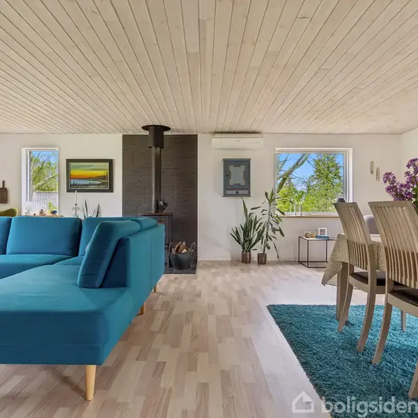 En stue med en blå hjørnesofa står på et trægulv. Et spisebord med seks stole er dækket af en blå tæppe. Rummet har lyse vægge, loftpaneler, og store vinduer.