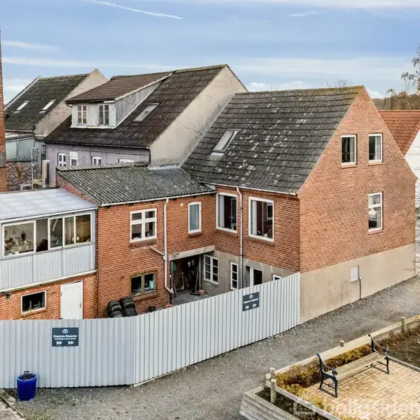 Bygning lavet af røde mursten, står ved siden af en høj skorsten. En metalindhegning omkranser bygningen, som har flere kvadratiske vinduer. Der er bænke og et bart træ i nærheden.