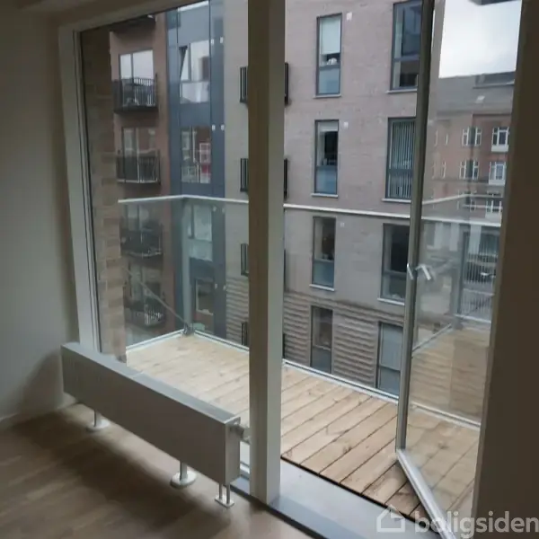 En glasdør fører ud til en lille altan. Indendørs vises et trægulv og en radiator. Udenfor ses en moderne bygning med flere vinduer.