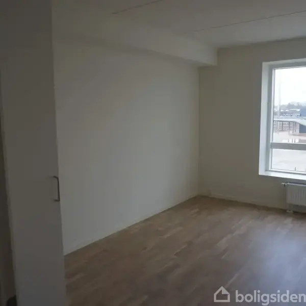 Et tomt værelse med trægulv, hvor et stort vindue lader lys strømme ind. I baggrunden ses en radiator under vinduet, og udenfor er der byudsigt.