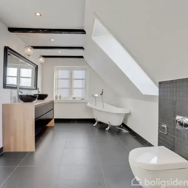 Et moderne badeværelse med fritstående badekar under et skråvindue. To håndvaske med stort spejl er på den anden væg. Rummet har sorte fliser og hvide vægge.