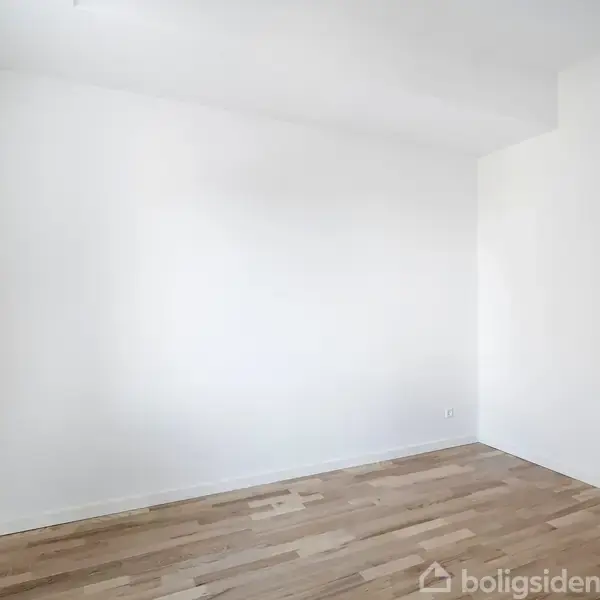 Et tomt rum har hvide vægge og lyst trægulv. Døren til venstre er halvt åben, hvilket skaber en minimalistisk atmosfære.