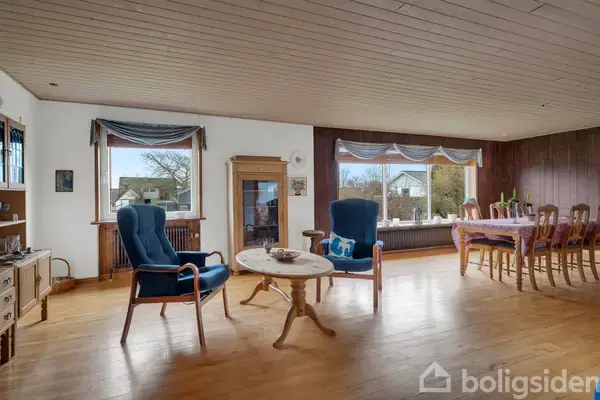 To blå lænestole står omkring et rundt sofabord i en stue med trægulv og store vinduer. I baggrunden ses et spisebord med stole og udsigt til en have.