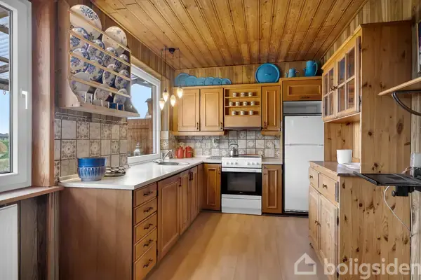 Køkken med træpaneler har skabslåger og skuffer i lys brun træfinish. Komfur og ovn står mellem skabe, og dekorative tallerkener er udstillet. Klinker dekorerer væggen. Lamper hænger fra loftet.