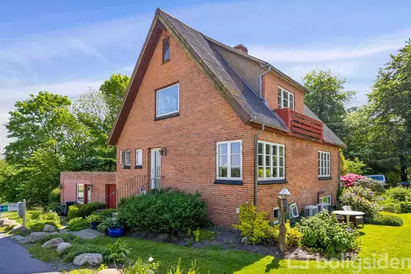 Hus i røde mursten med skråt tag og hvide vinduer, omgivet af grønne buske og blomster i en velplejet have. Blå himmel og frodige træer danner baggrunden.