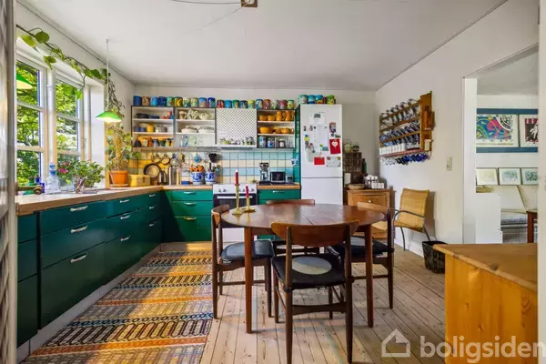 Et rundt spisebord med træstole står i et køkken med grønne kabinetter og farverige krus på hylder. Rummet er lyst med planter i vindueskarmen og vægkunst.