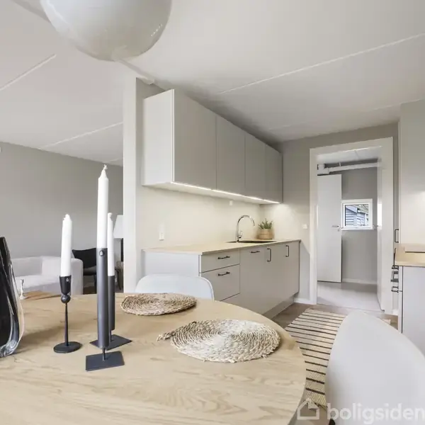 Et rundt spisebord med stearinlys og en vase står i en moderne, lys stue-køkken. Funktionelle køkkenskabe med indbygget belysning ses i baggrunden nær en åben dør.