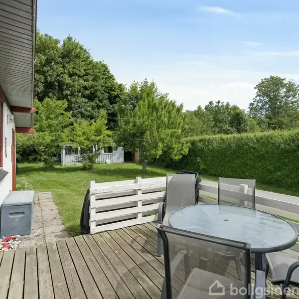 Terrasse med glasbord og stole står på trædæk; begrænset af hegn og grøn græsplæne i baggrunden med høje træer. Huset har en hvid og rød facade.