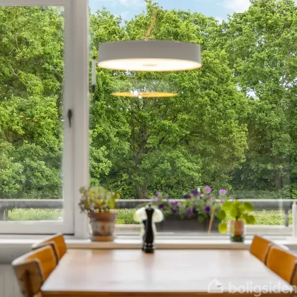 Et spisebord står stille foran et stort vindue, hvor man ser grønne træer udenfor. Bordet har fire stole og er pyntet med planter i potter.