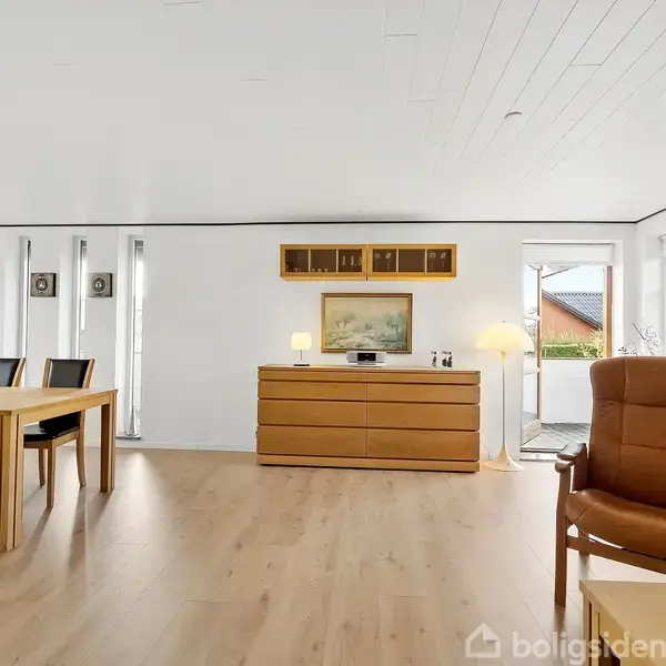 Et spisebord med stole står i en lys stue. En kommode med lamper og billede hænger på væggen. Store vinduer med udsigt omgiver rummet.