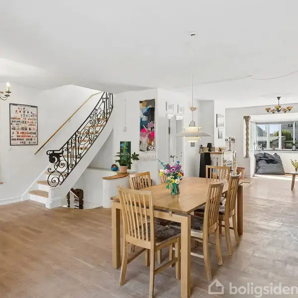 Spisestue med træbord og stole, dekoreret med blomster, i moderne hjem. Hvidmalede vægge, trappe med smedejernsgelænder og verdenskort. Opholdsområde i baggrunden med sofaer og store vinduer.